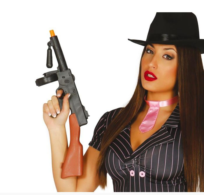 A woman wearing a black fedora hat, a black pinstripe blouse, and a shiny pink necktie accessory, holding a toy submachine gun prop against a white background.