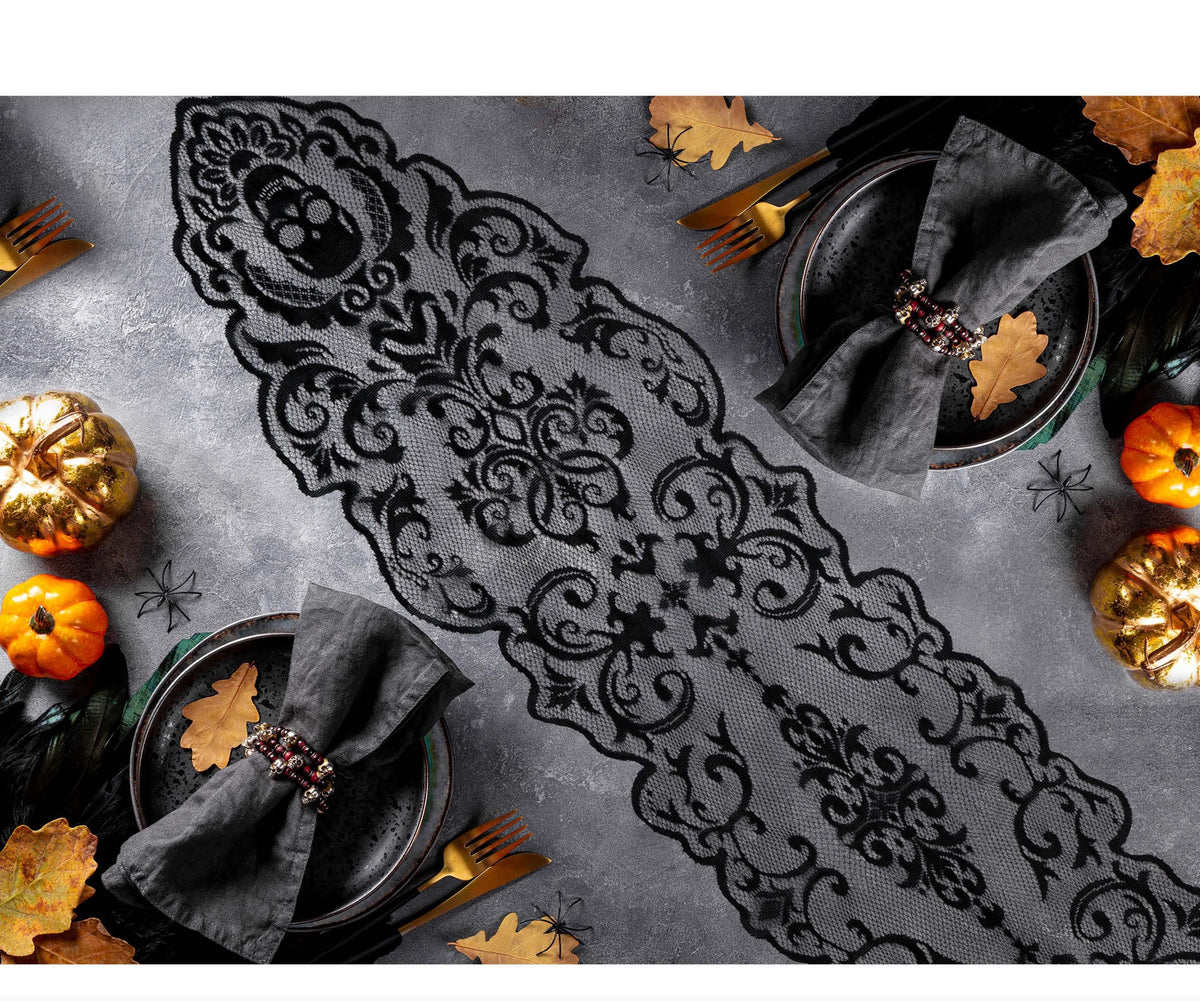 A black lace table runner with an ornate gothic damask pattern laid diagonally across a gray textured tabletop. The scene is styled with dark gray napkins, gold forks, small orange pumpkins, and autumn leaves, with two plates partially visible.