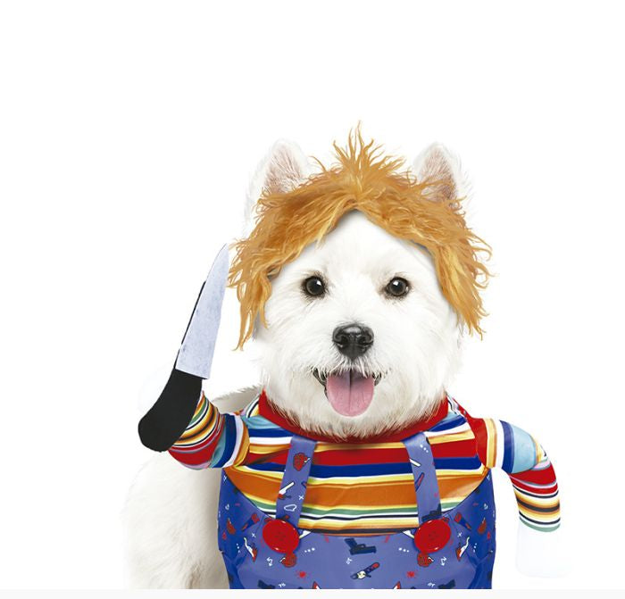 A white dog dressed as Chucky, wearing a blonde wig, a colorful striped shirt, blue overalls with red splatter details, and holding a toy knife, photographed against a plain white background.