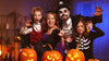 A family of four posing in Halloween costumes in a spooky, decorated setting with jack-o'-lanterns. The father wears a black pirate costume with a top hat and skull-and-crossbones emblem, the mother wears a purple dress, the boy wears a red and white vampire outfit, and the girl wears a black skeleton costume.