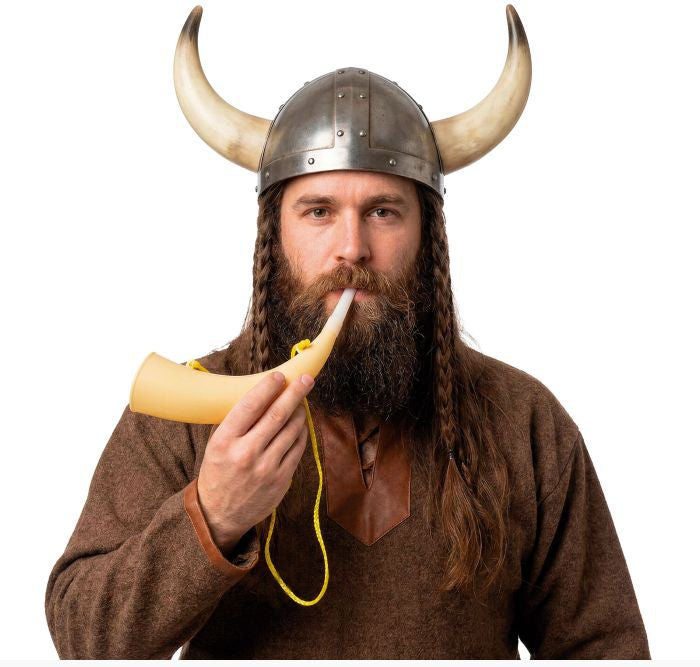 A man wearing a Viking-style horned helmet and brown tunic holds a small horn flute to his mouth against a plain white background. 