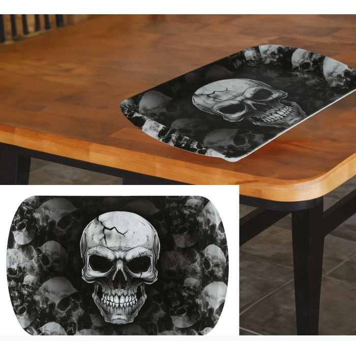 A rectangular black and white skull-patterned tray is shown resting on a wooden dining table, with a smaller inset close-up of the tray design below. The design features a large central skull over a background of smaller skulls in grayscale.