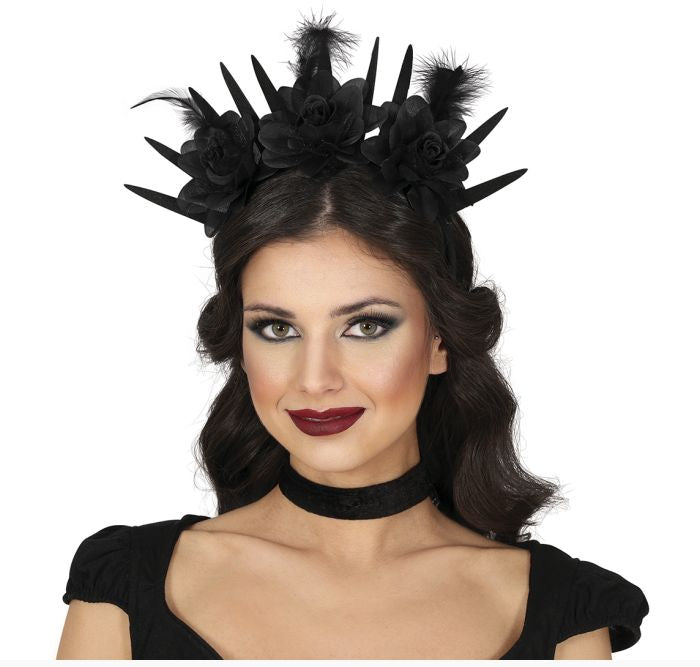 A woman modeling a black gothic-style fascinator headpiece with artificial flowers, spikes, and feathers, paired with a black choker and black top against a white background.