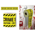 Yellow and black caution-style toilet paper tape shown in two product views: a roll wrapped around a toilet paper holder on a white wall with red blood-splatter styling, plus a separate roll laid flat. The tape reads 'CRIME SCENE - DO NOT CROSS' with black text on a bright yellow background.
