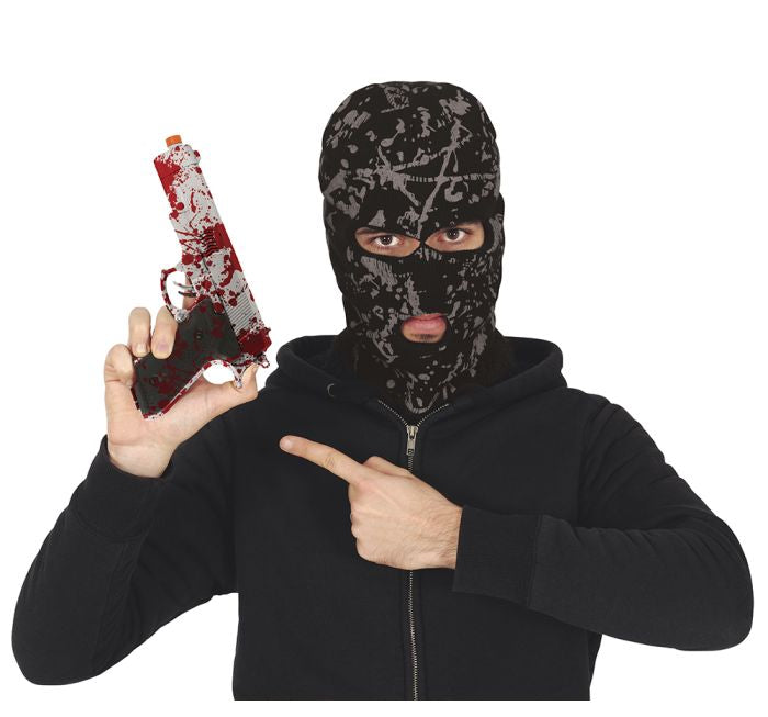 A person wearing a black and gray patterned balaclava mask and a black zip-up hoodie, holding a toy gun splattered with red fake blood and pointing at it. The background is solid white, with a copyright watermark for Fiestas Guirca at the bottom.
