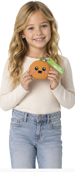 A young girl with long blonde hair smiles while holding a small orange plush character toy with a green 'WELCOME' tag. She is wearing a cream ribbed long-sleeve top and light blue jeans against a plain white background.