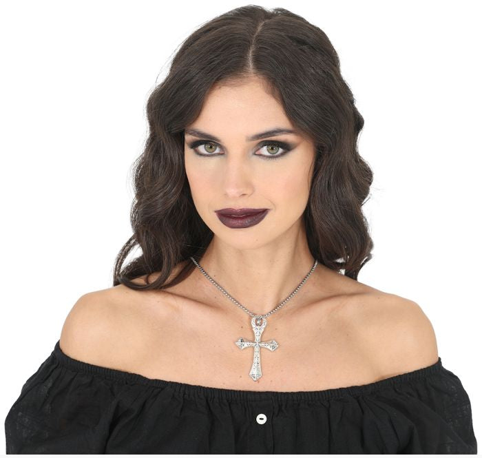 A woman with dark wavy hair and dark lipstick wears a silver cross pendant necklace with clear crystals, paired with a black off-the-shoulder top against a white background.