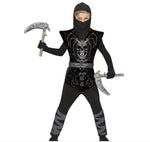 A child modeling a black ninja costume with a skull graphic on the chest, holding toy scimitar and scythe props against a white background.