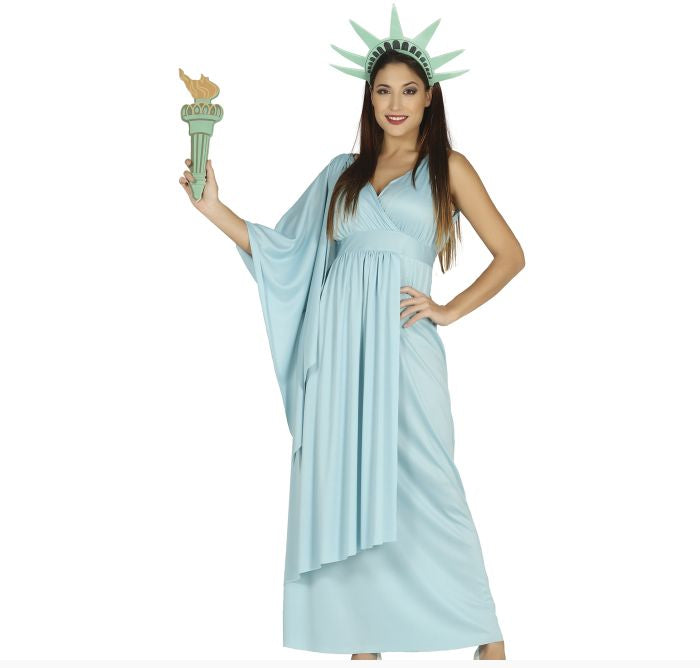 A woman modeling a light blue Statue costume against a plain white background, holding a matching green and gold toy torch. The costume includes a long flowing dress and a green crown with spikes.